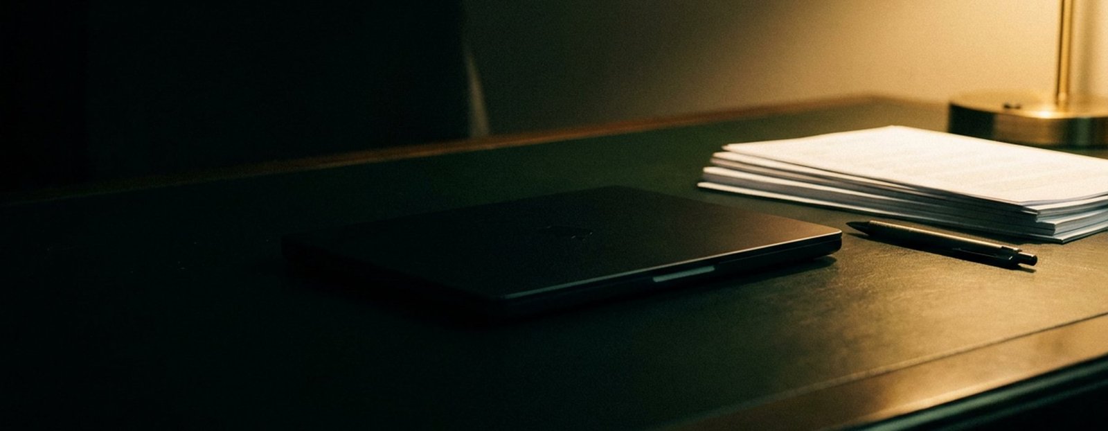 Mesa de trabalho em plano rasante: notebook fechado, documentos e a luz quente da luminária